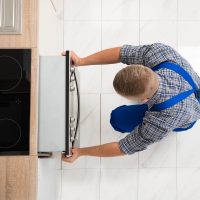 High Angle View Of Man In Overall Repairing Oven In Kitchen